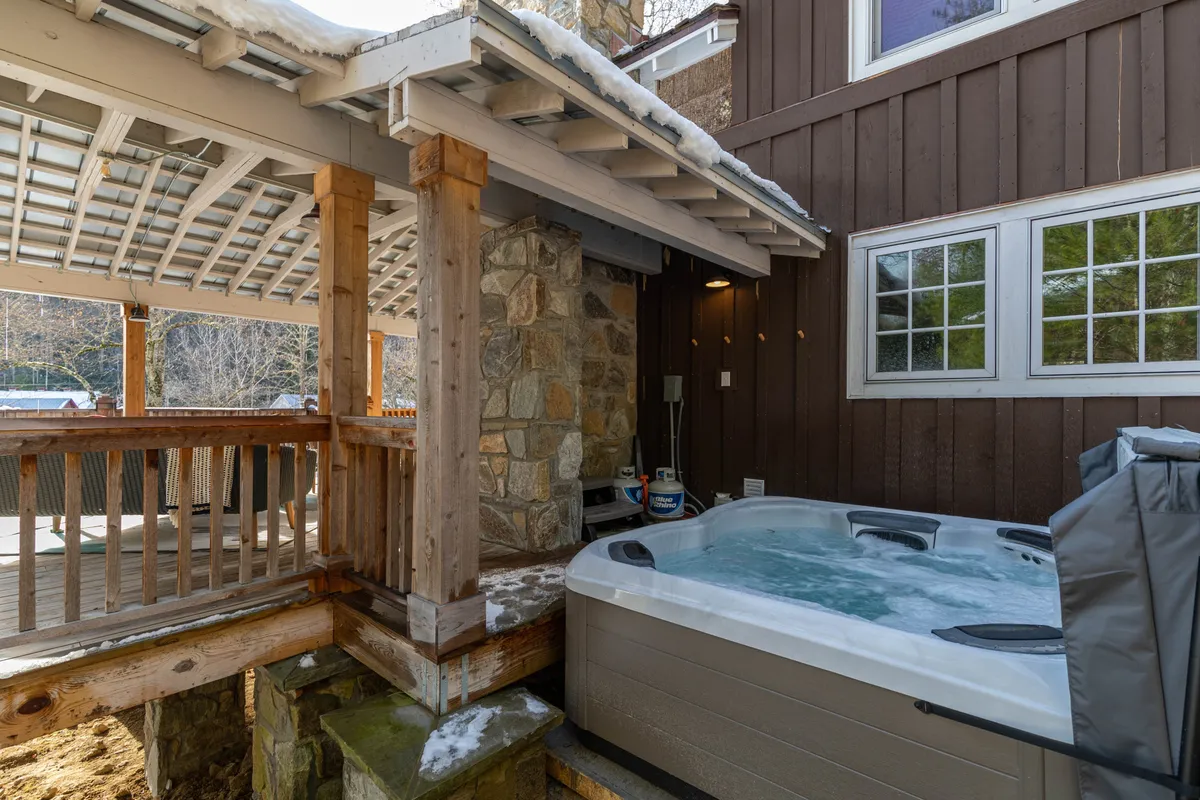 This image features a private outdoor hot tub set on a wooden deck with a stone wall and overhead pergola, surrounded by lush natural greenery. The scene exudes tranquility and invites guests to soak and unwind while enjoying serene views.