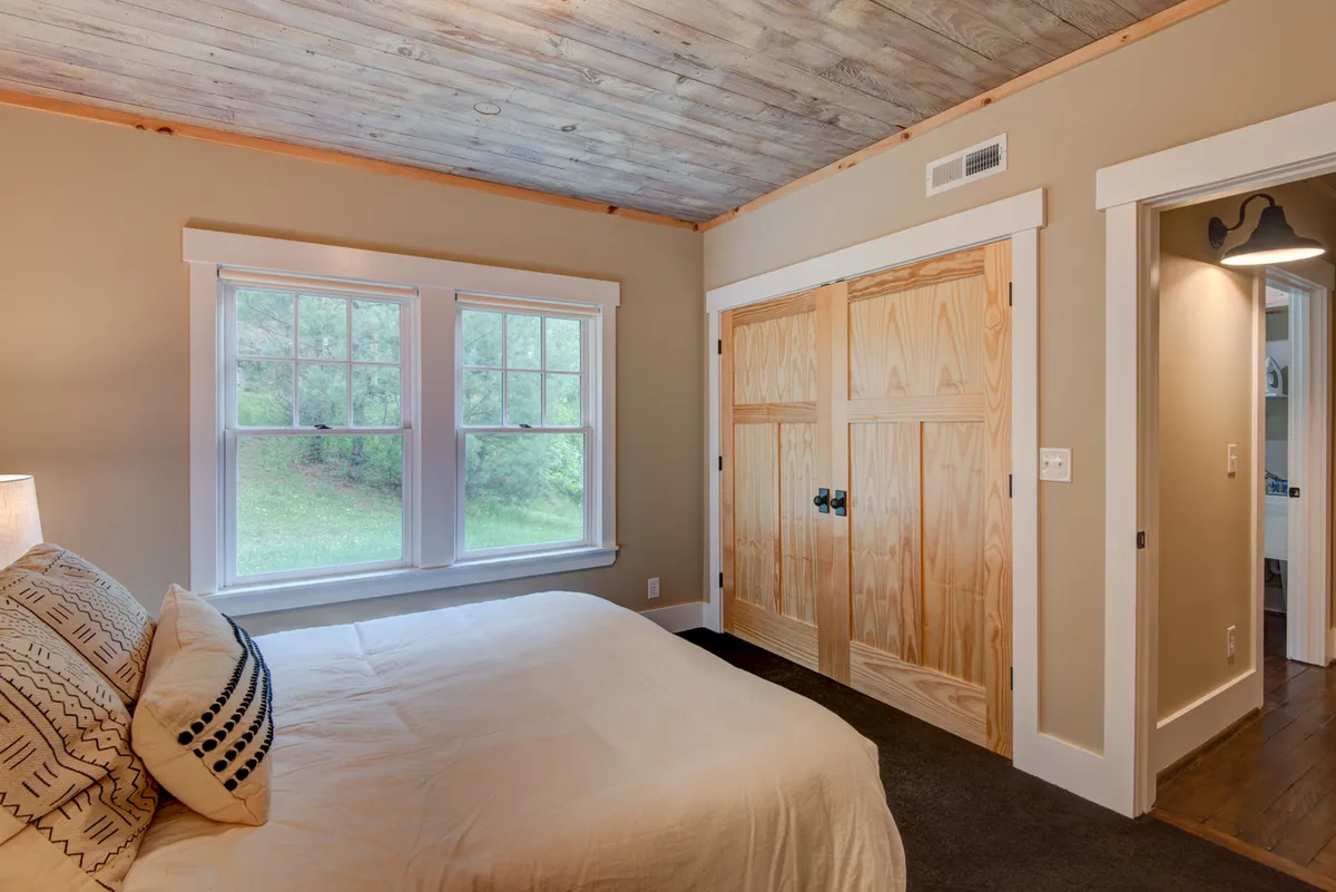 A tranquil bedroom showcasing a bed dressed in luxurious decorative pillows and plush linens, bathed in natural light from large windows and complemented by warm wooden closet doors.