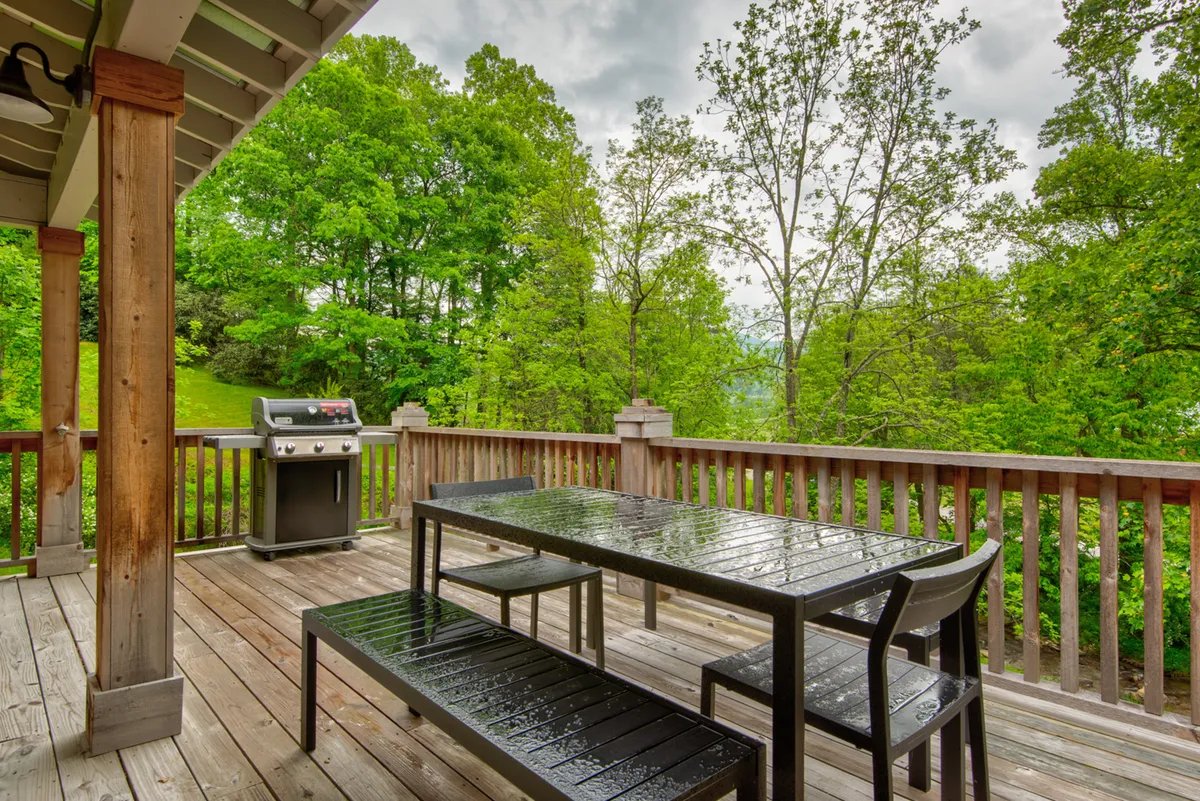 A wooden deck area set up for alfresco dining, with a table and chairs, grill nearby, and green foliage all around. The deck offers a peaceful view, perfect for meals and social gatherings outdoors.