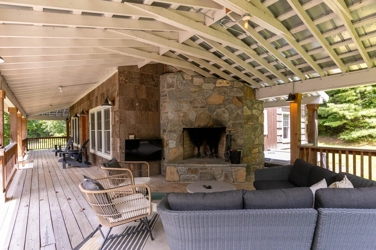 A spacious covered deck featuring a stunning stone fireplace, comfortable outdoor seating, exposed wooden beams, and lush natural surroundings. The setting is ideal for relaxation or gathering with family, with ample room for outdoor entertainment and dining.