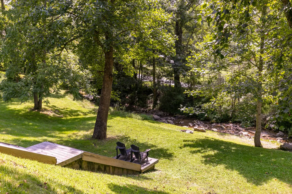 Two black chairs positioned on lush grass beside a gently flowing creek, framed by tall trees. The serene setting offers a quiet spot for relaxation, with nature close at hand.