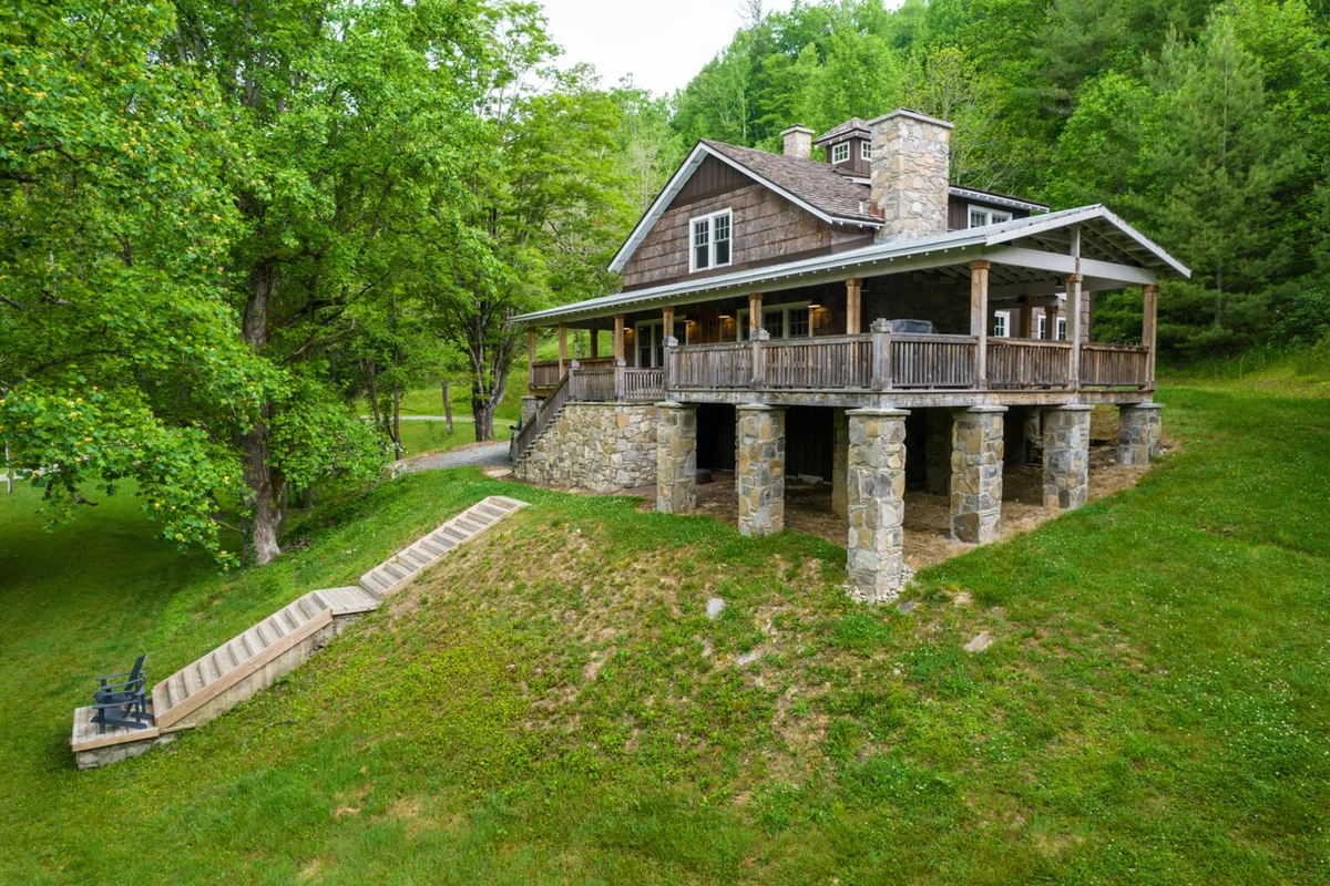 A wide exterior shot of Orchard House’s generous porch supported by stately stone pillars, with an expansive deck overlooking the surrounding greenery. The scene is peaceful and inviting, perfect for evening gatherings, watching sunsets, or exploring the natural landscape just steps away.