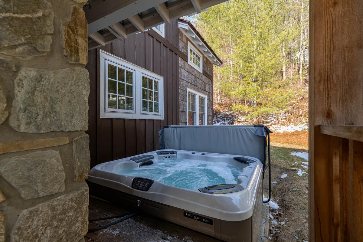A bubbling hot tub set against a stone wall under a covered porch, with tranquil outdoor views visible from the tub. The scene feels private and serene, inviting relaxation after a day of adventure.