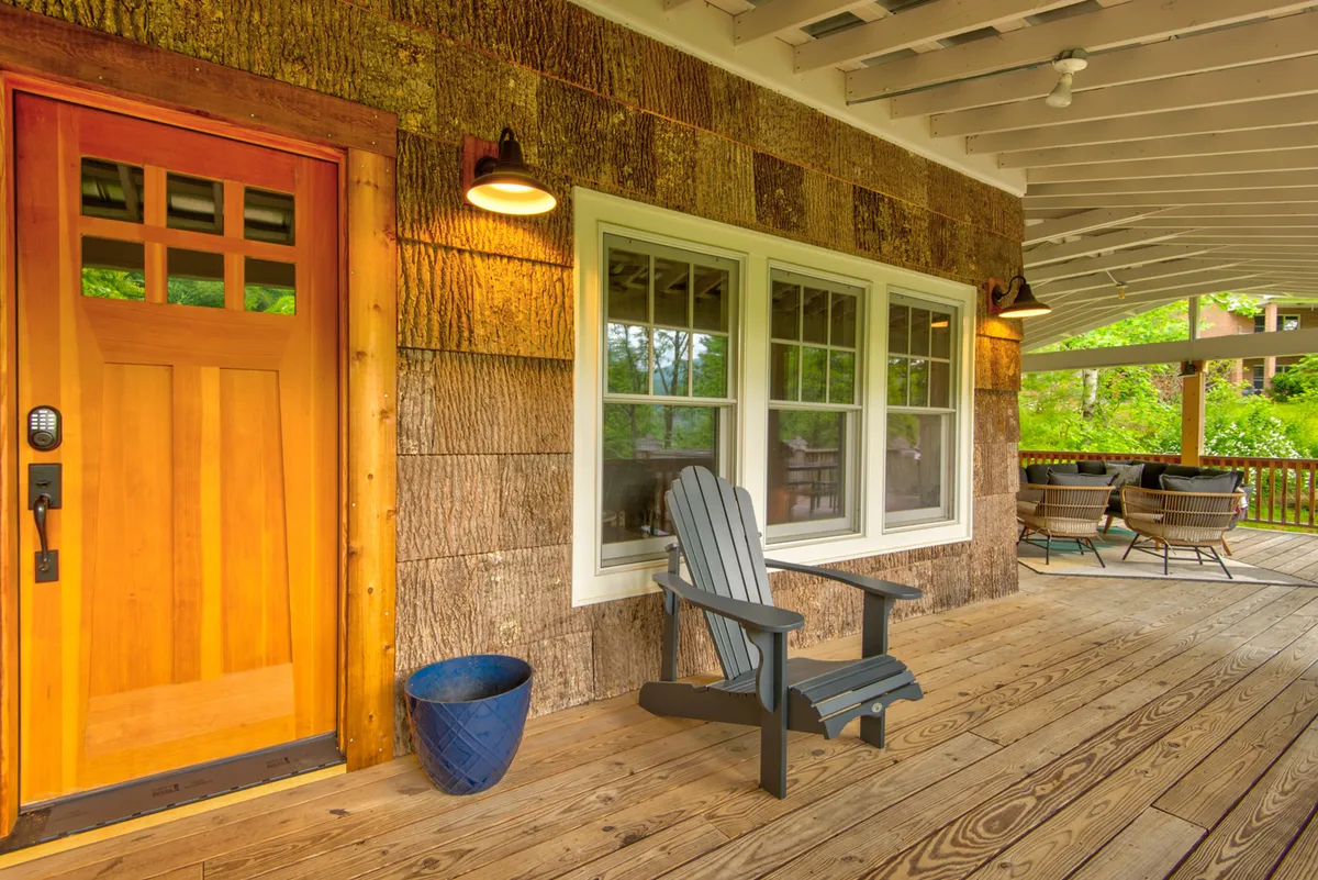 This exterior image features the front door of Orchard House, a wooden chair, a decorative plant pot, and prominent windows. The inviting porch setting hints at a welcoming, homey atmosphere and subtly suggests the house's connection with the outdoors.