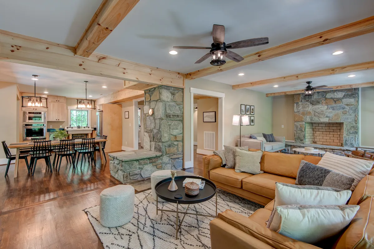 This image captures a warmly lit, inviting living room featuring a striking stone fireplace, exposed wooden beams, a comfortable sofa, and a view toward the adjacent dining area. The space feels perfect for both intimate family gatherings and larger social occasions, with clear sightlines between the sitting and dining zones.