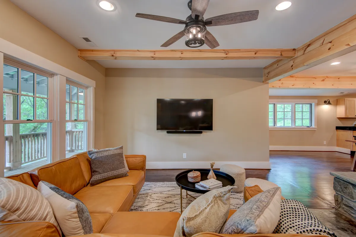 A welcoming, sunlit living room with a plush leather sofa, a stylish coffee table adorned with tasteful decor, a ceiling fan, and expansive windows letting in natural light. The space feels modern yet inviting, perfect for relaxing, socializing, or unwinding with digital entertainment.