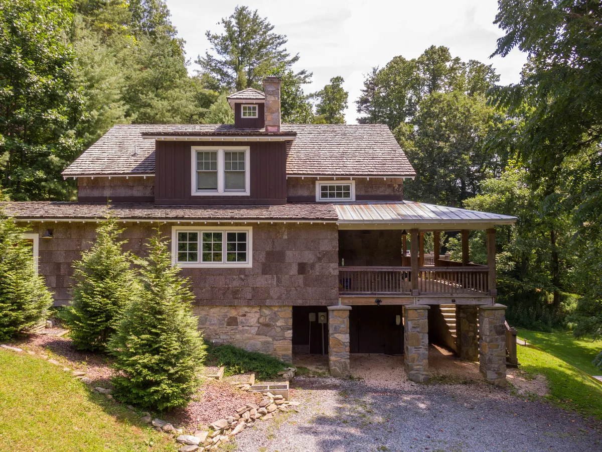 The exterior of the home shows a sturdy stone foundation, wooden shingles, and a welcoming deck area, all surrounded by mature trees. The scene hints at outdoor gatherings, relaxing mornings, and the seamless blend between comfy living and the great outdoors.