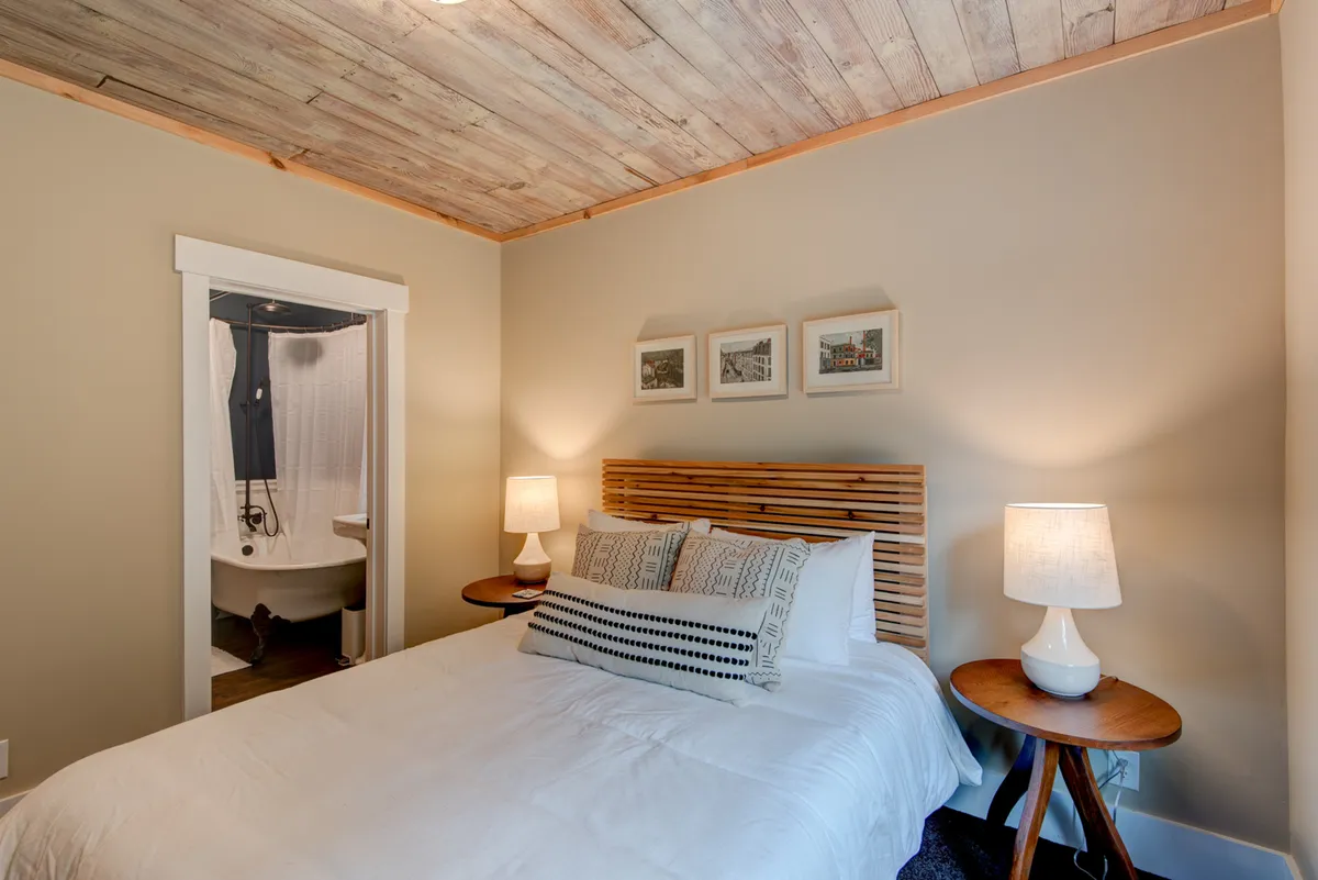 A serene bedroom featuring a comfortable bed with plush bedding, wall art, bedside lamps, and a classic clawfoot bathtub visible in the corner. The gentle lighting creates a cozy, restful atmosphere perfect for unwinding.