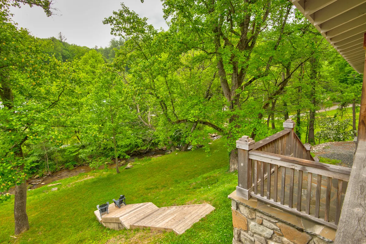 A peaceful outdoor seating area set on a wooden deck overlooks a lush green landscape dotted with tall trees. A clear stream runs nearby, inviting visitors to relax by the water or observe local wildlife. The space feels secluded and immersed in nature, perfect for enjoying the sights and sounds of the Blue Ridge outdoors.
