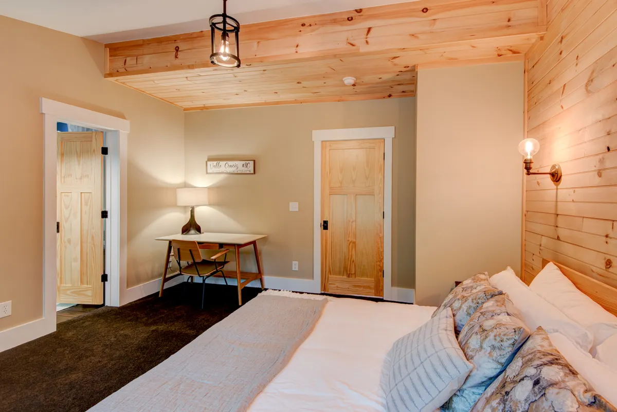 A welcoming bedroom with a bed adorned with decorative pillows, a wooden desk with a lamp for work or study, and wooden doors. Natural light streams in, making the space inviting and functional.