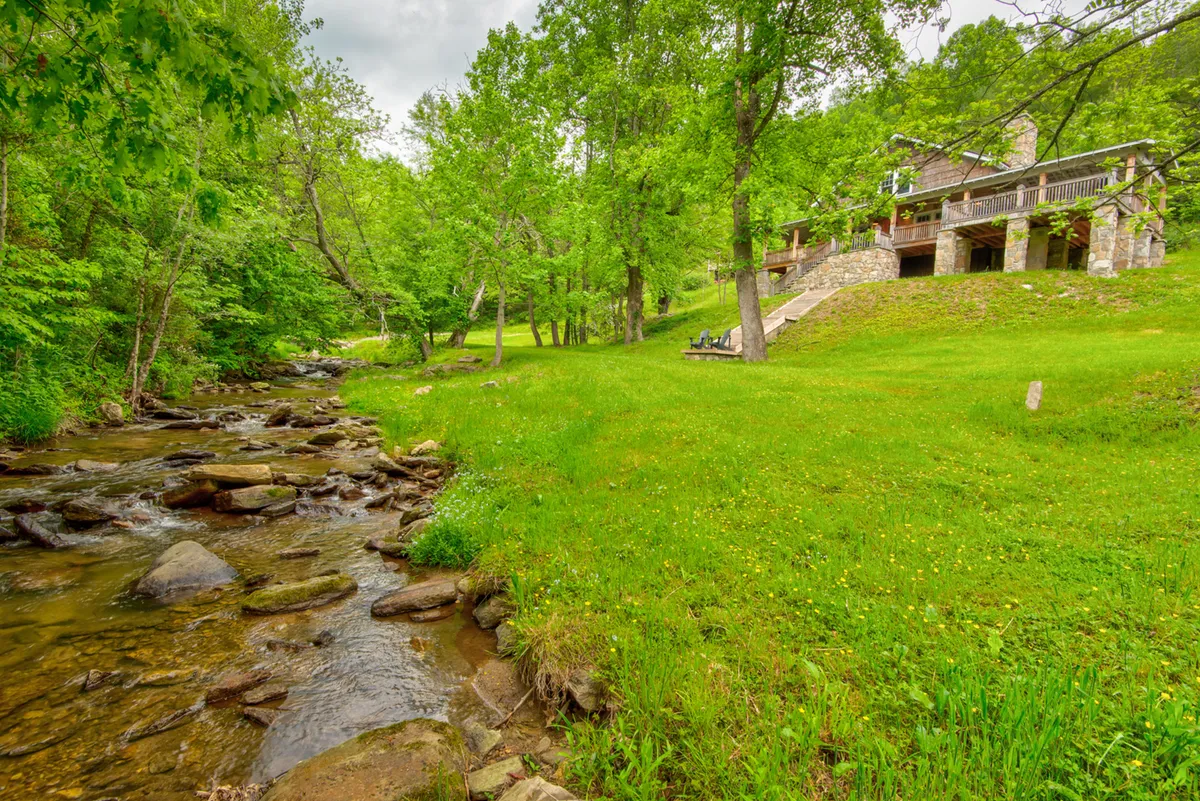This exterior image features the stone house nestled beside Crab Orchard Creek, with lush greenery surrounding the gentle stream. The scene invites viewers to imagine relaxing or picnicking by the water, with easy access to nature right in the front yard.