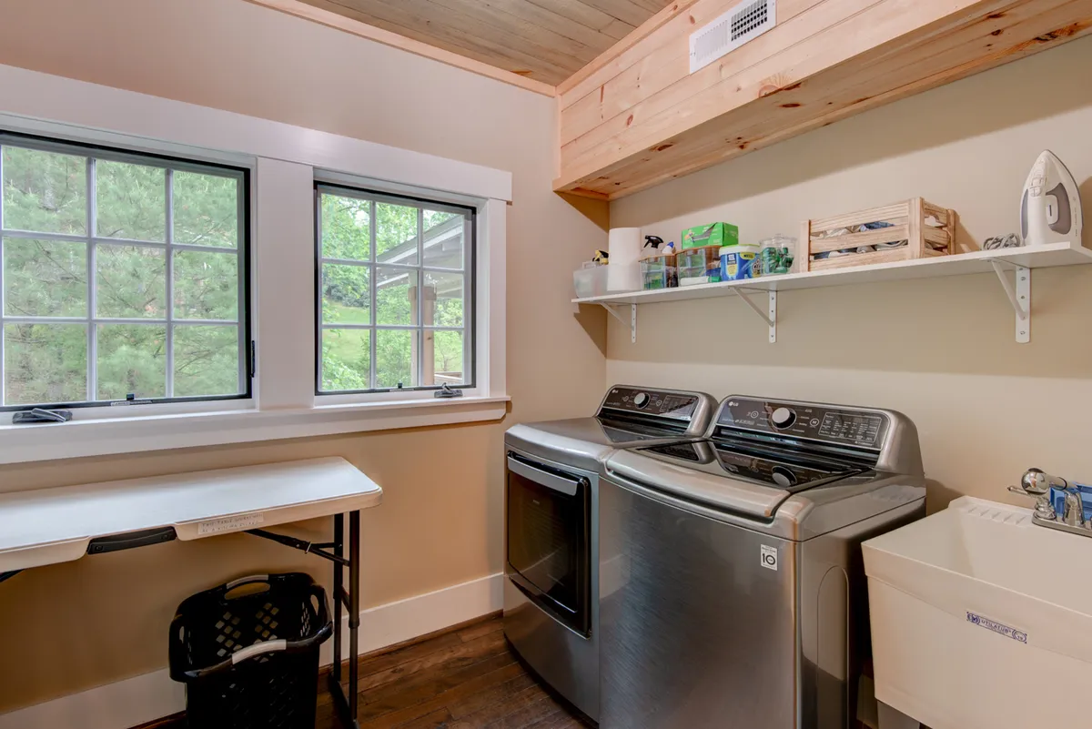 A bright, functional laundry area featuring modern washing machines, a utility sink, and organized shelves beneath large sunlit windows. The space feels clean and efficient, reflecting the home's well-stocked essentials and guest convenience.