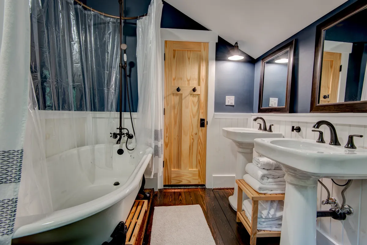 A stylish bathroom featuring a freestanding clawfoot bathtub, double pedestal sinks, wooden door, and large mirrors. The space is elegant and spacious, perfect for relaxing and self-care.
