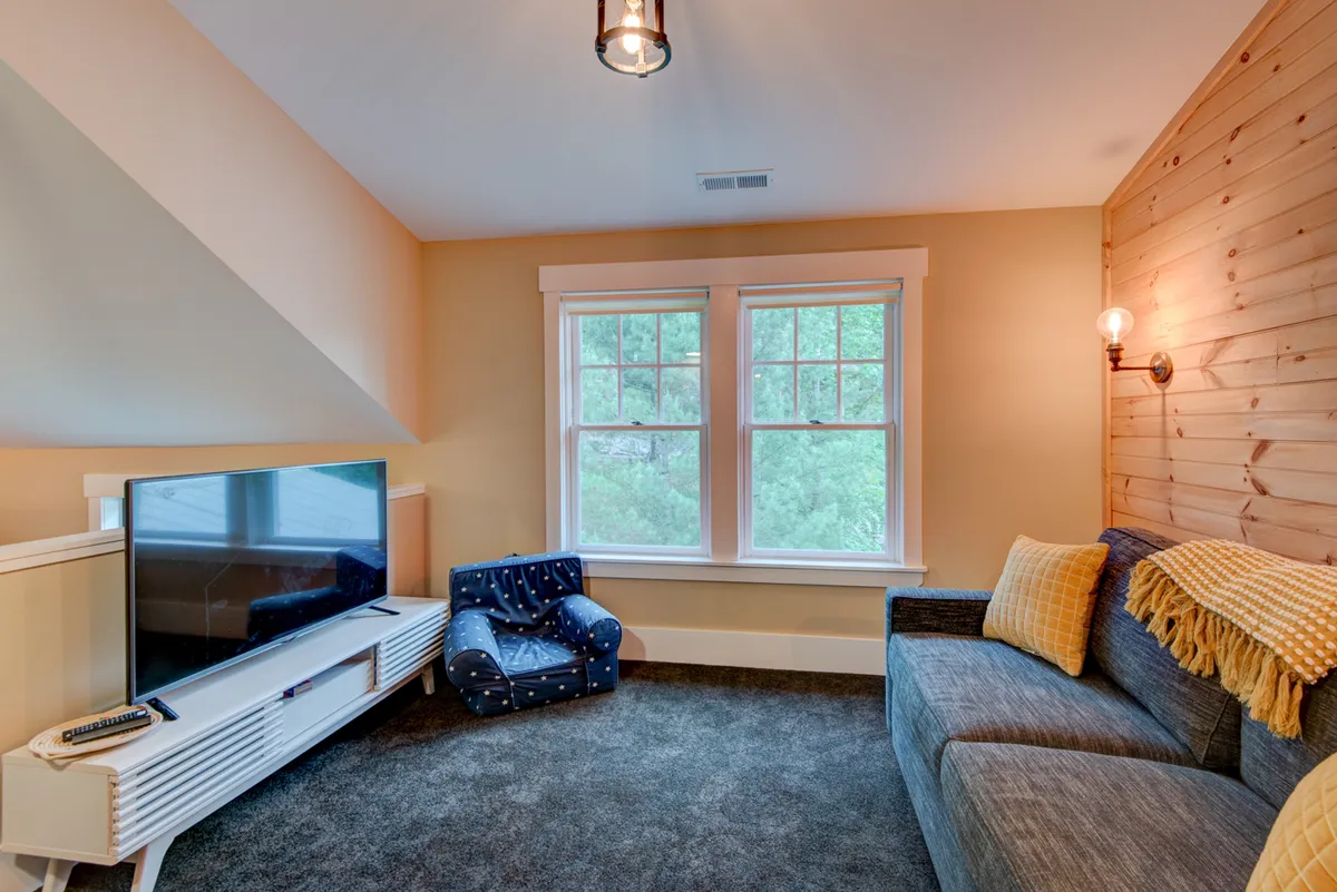A comfortable media and lounge area is shown here, with a smart TV, plush seating, a wood-paneled accent wall, and large windows letting in natural light. The arrangement is ideal for watching movies, playing games, or simply relaxing with family and friends.