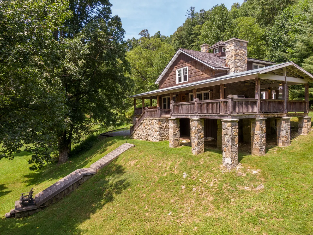 The image features the welcoming exterior of Orchard House, highlighted by its charming stone foundation and inviting porch with wooden railings. Surrounded by lush greenery and mature trees, the house is set in a tranquil, natural landscape. The porch offers a perfect spot for relaxing, taking in sunset views, and enjoying the soothing sounds of the nearby creek.